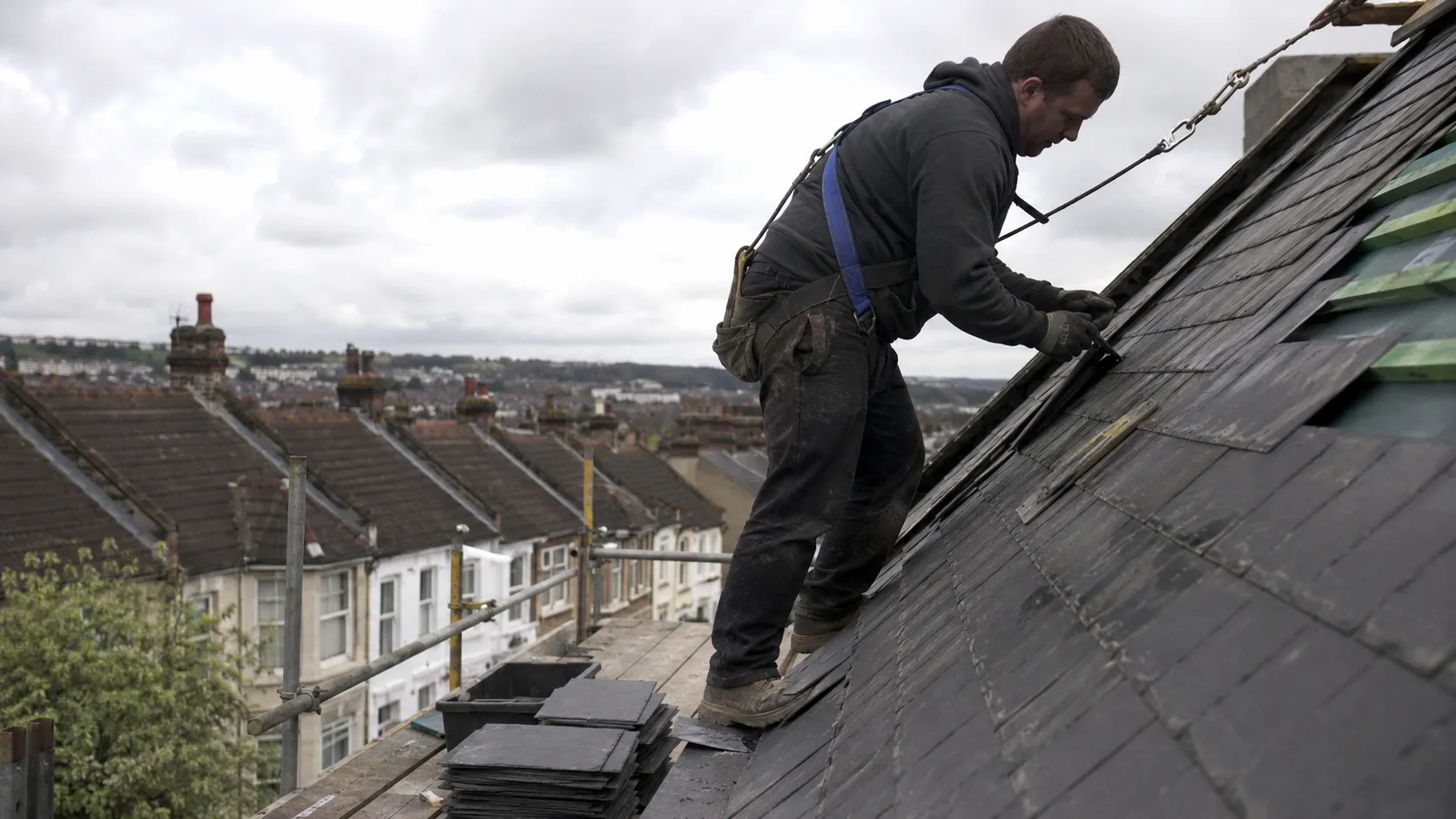 A Ridgeline roofer, harnessed, laying natural slate on a Sheffield rooftop with terraced streets and hills behind